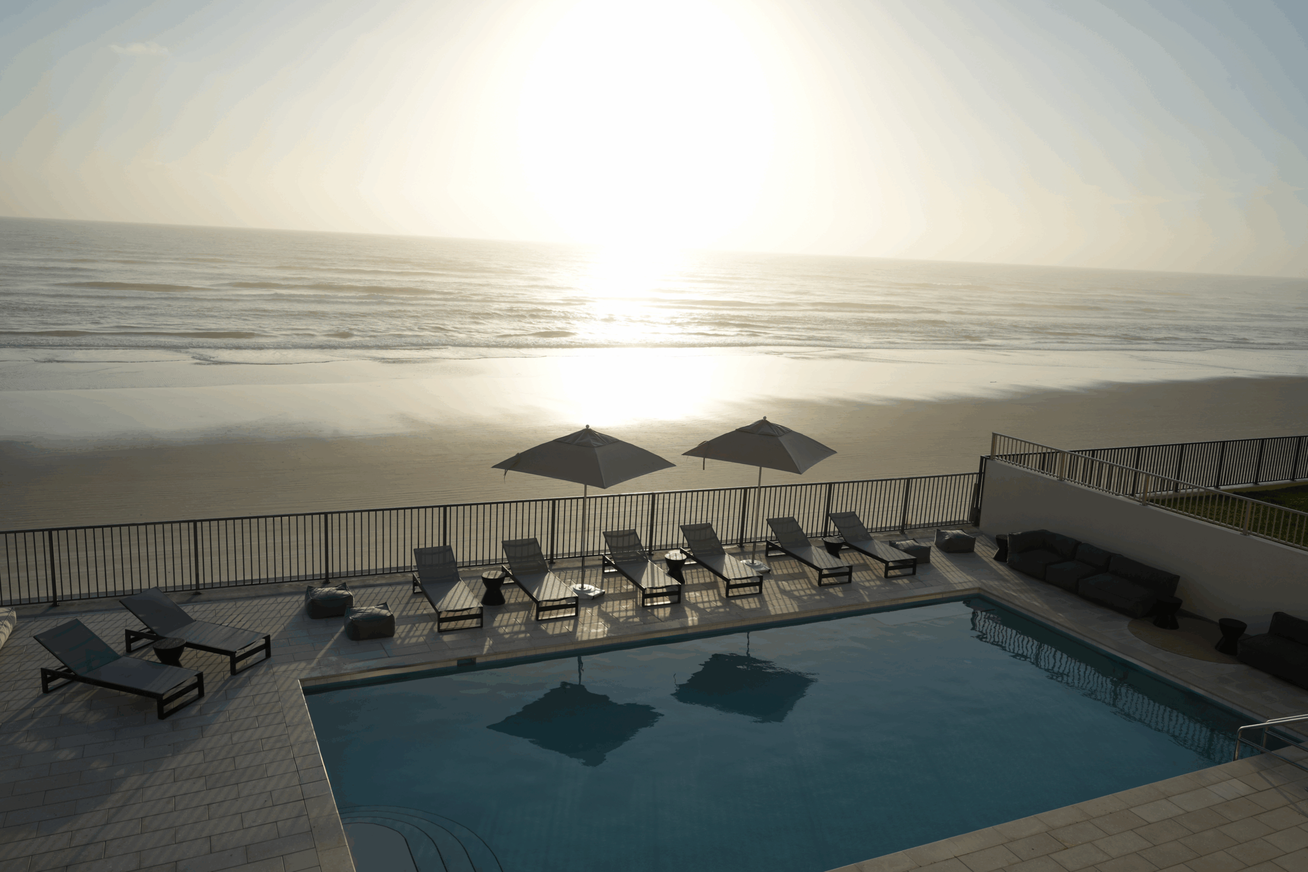 Beachfront pool at Ambros Daytona Hotel with lounge chairs and umbrellas overlooking the Atlantic Ocean at sunset