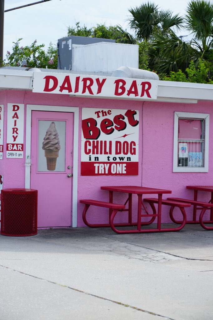 Front view of the Dairy Bar in Daytona Beach featuring bright pink walls, red signage, picnic seating, and a vintage soft-serve cone decal.