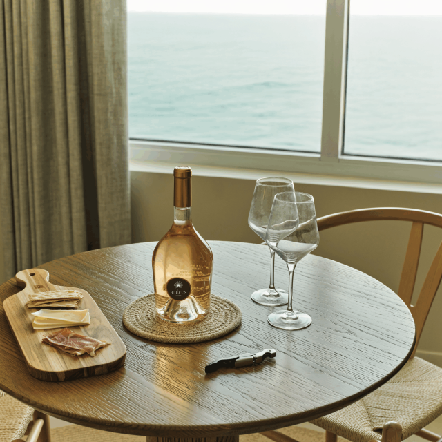 Bottle of Ambros rosé wine, two glasses, and a charcuterie board on a wooden table with ocean views.