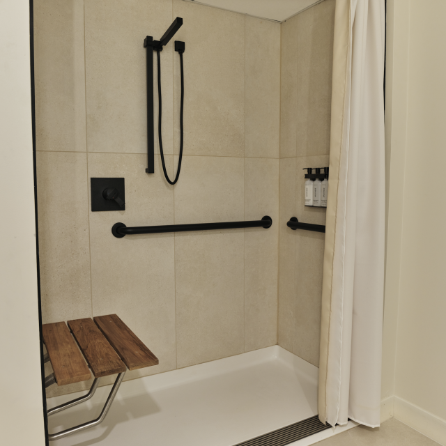 ADA-accessible roll-in shower at Ambros Hotel with teak bench, grab bars, matte black fixtures, and wall-mounted toiletries